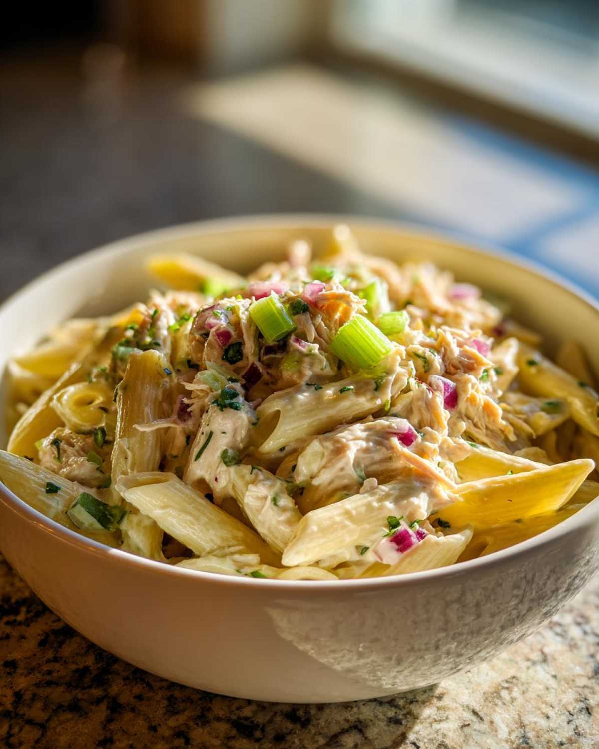 Close-up of creamy Tuna pasta salad featuring penne, tuna, red onion, and celery in a white bowl.
