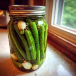 A glass jar filled with bright green dilly beans, garlic cloves, and spices, ready for pickling.