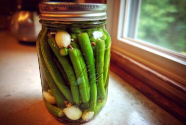 A glass jar filled with bright green dilly beans, garlic cloves, and spices, ready for pickling.