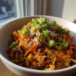 A close-up of a serving of egg roll in a bowl, featuring ground meat, shredded cabbage, and carrots, topped with sesame seeds and fresh green onions.