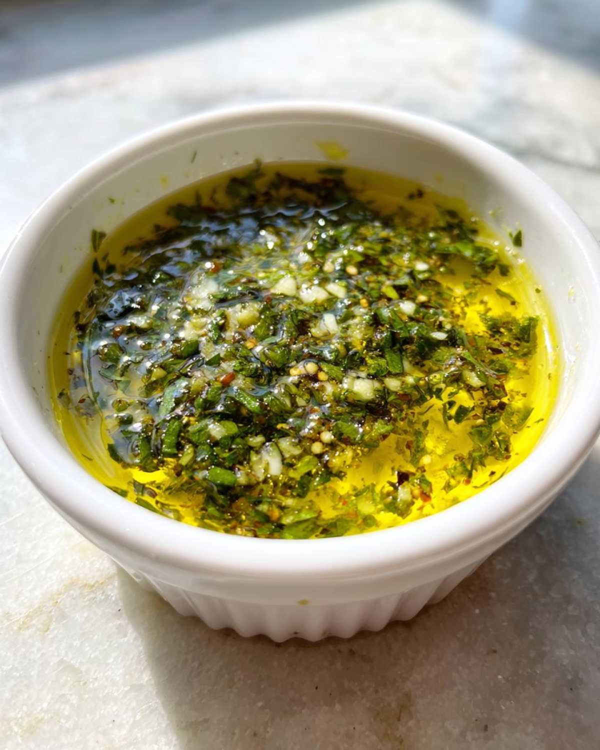 Close-up of a white ramekin filled with vibrant olive oil bread dip, featuring chopped herbs and garlic floating in golden oil.