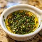 Close-up of a white dipping bowl filled with vibrant olive oil bread dip featuring chopped green herbs and spices.