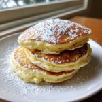 A tall stack of three fluffy buttermilk pancakes dusted generously with white powdered sugar on a white plate.