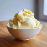 A generous serving of creamy, fluffy mashed potatoes piled high in a white bowl on a wooden surface.