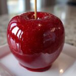 A single, perfectly coated, glossy red candy apple sitting on a white square plate.