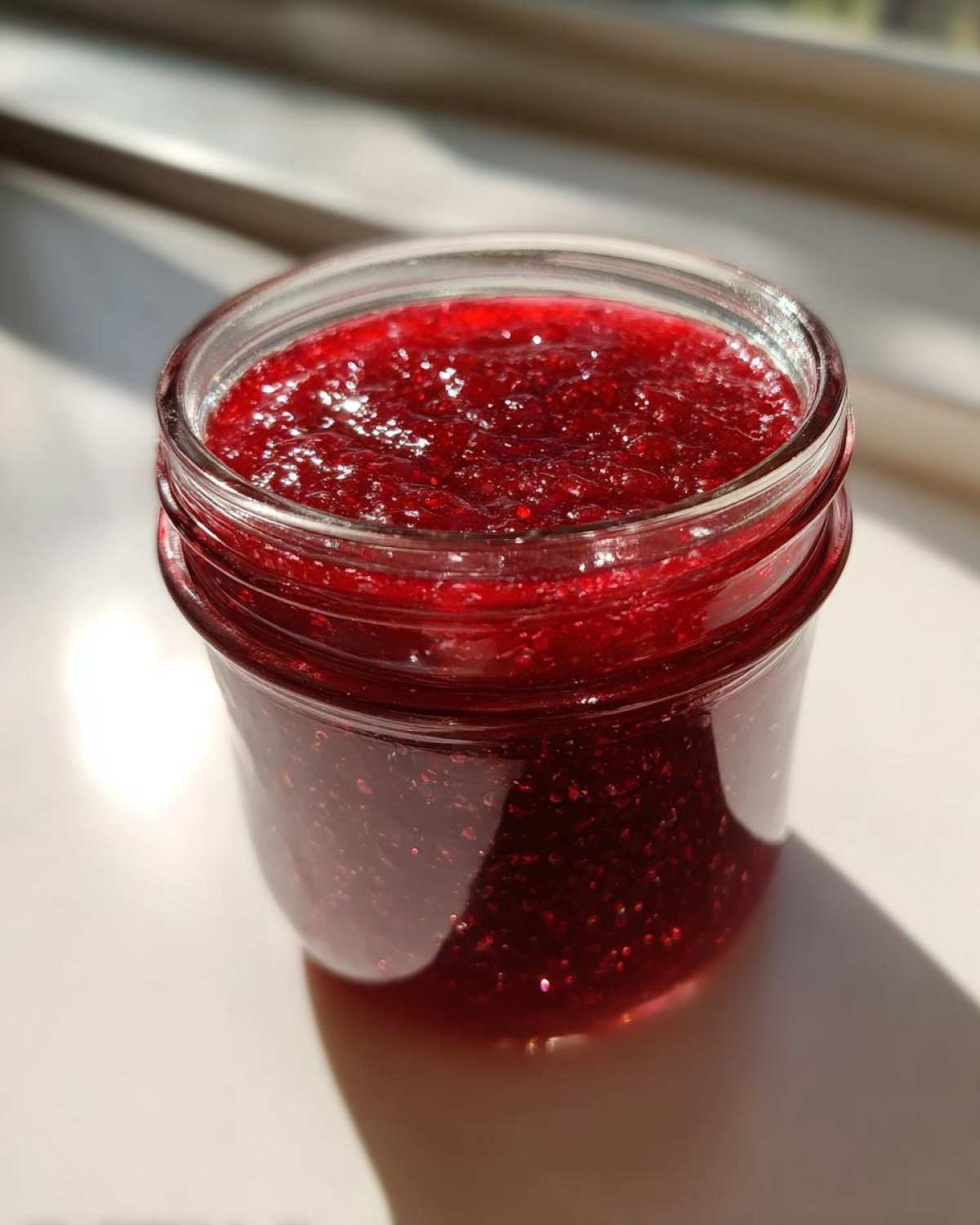 Close-up of a small glass jar filled with vibrant, glistening homemade raspberry jam, catching the sunlight.