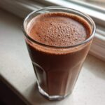 Close-up of a tall glass filled with rich, frothy homemade chocolate milk sitting on a light windowsill.
