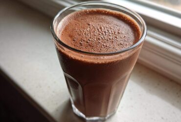 Close-up of a tall glass filled with rich, frothy homemade chocolate milk sitting on a light windowsill.