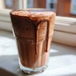 A tall glass filled with thick, frothy frozen hot chocolate, with chocolate syrup dripping down the side.