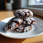 A stack of four fudgy chocolate crinkle cookies heavily dusted with white powdered sugar on a white plate.