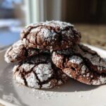 A stack of four fudgy chocolate crinkle cookies dusted heavily with powdered sugar, sitting on a white plate.
