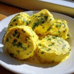 Four scoops of bright yellow homemade garlic butter mixed with green herbs served on a white plate.