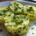 Four chilled rounds of homemade garlic butter, richly mixed with fresh green herbs, sitting on a white plate.