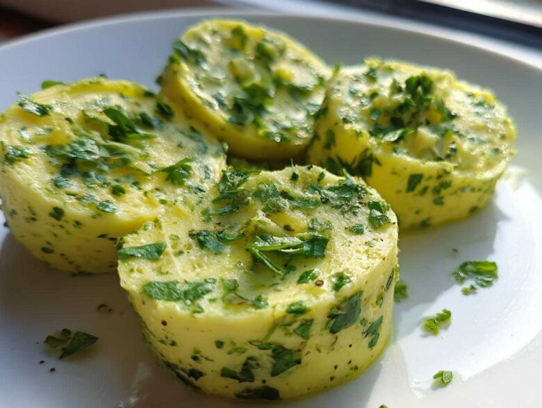 Four chilled rounds of homemade garlic butter, richly mixed with fresh green herbs, sitting on a white plate.