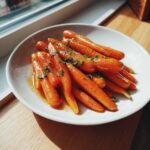 A white bowl filled with glossy, glazed roasted carrots, sprinkled with fresh parsley, sitting on a wooden surface near a window.