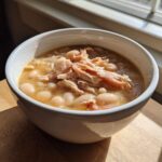 Close-up of a white bowl filled with ham and white bean soup, topped with shredded ham.