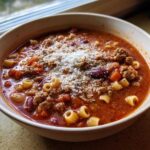 A close-up of a steaming bowl of rich, tomato-based pasta e fagioli topped with ground meat, beans, and grated Parmesan cheese.