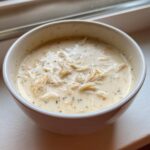 A close-up of a white bowl filled with creamy homemade cream of chicken soup featuring visible shreds of chicken and herbs.