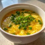Close-up of a steaming bowl of bright yellow egg drop soup garnished generously with fresh green scallions.
