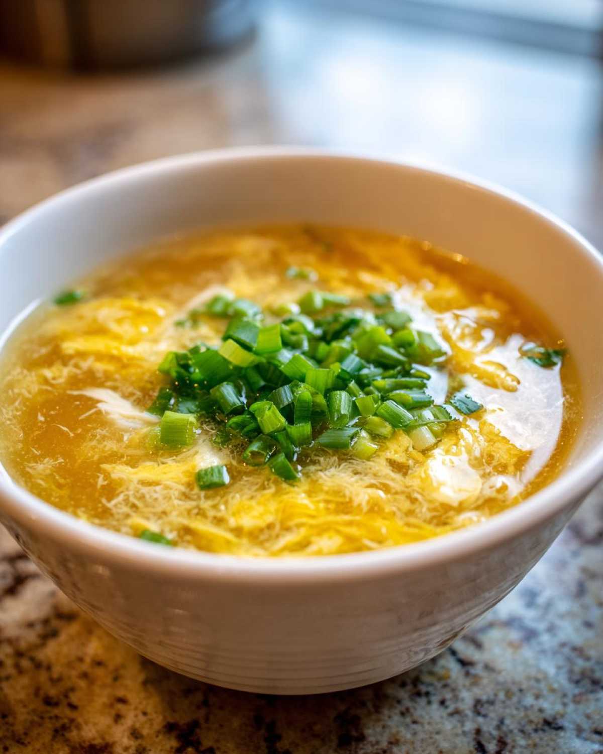 A close-up of a white bowl filled with savory, golden egg drop soup topped with fresh chopped green onions.