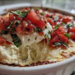 A spoonful of hot, cheesy bruschetta dip being lifted from a white baking dish, topped with fresh tomatoes and basil.