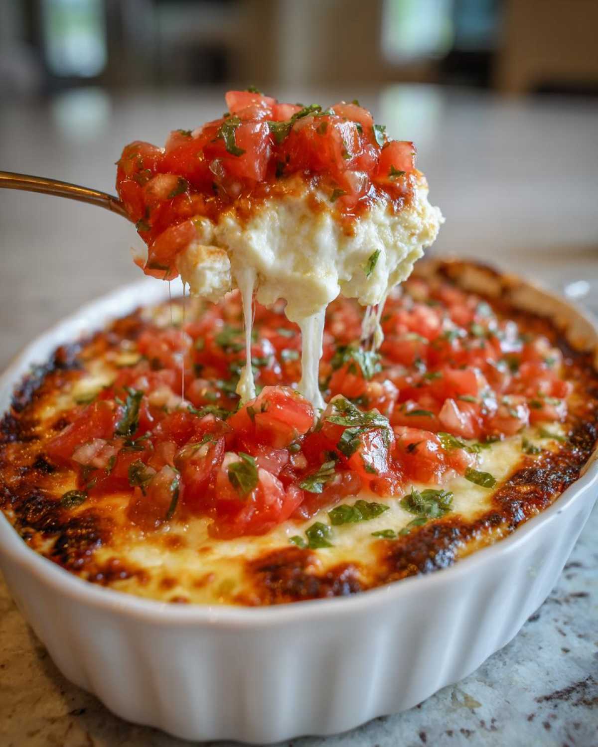 A spoonful of hot, cheesy bruschetta dip being lifted from a white baking dish, showing long cheese pulls and fresh tomato topping.