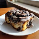 A single, gooey chocolate cinnamon roll topped with white icing, sitting on a white plate.