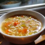 A close-up of a white bowl filled with hot Italian penicillin soup featuring broth, orzo pasta, carrots, and celery.