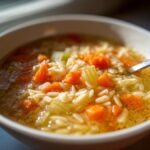 A close-up view of a white bowl filled with hot Italian penicillin soup, featuring orzo pasta, diced carrots, and celery in a clear broth.
