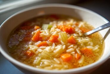 A close-up view of a white bowl filled with hot Italian penicillin soup, featuring orzo pasta, diced carrots, and celery in a clear broth.
