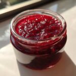 Close-up of a glass jar filled with glistening, deep red homemade raspberry jam, catching bright sunlight.