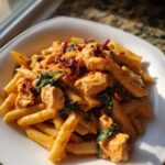 A close-up of a bowl of Marry Me Chicken Pasta featuring penne, chicken chunks, spinach, and sun-dried tomatoes in a creamy sauce.