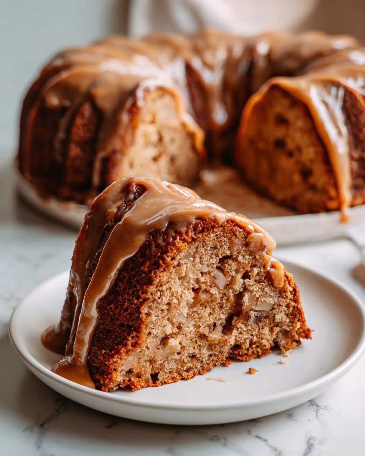 A close-up slice of moist apple bundt cake generously drizzled with caramel sauce, with the rest of the cake in the background.