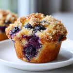 A close-up of a moist blueberry muffin topped with a sugary crumb topping, bursting with fresh blueberries.