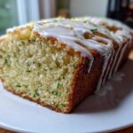 A loaf of moist lemon zucchini bread, sliced to show green zucchini shreds, topped with a thick white lemon glaze and zest.