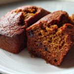 Close-up of two rich, dark brown mini banana bread loaves, one broken open to show the moist crumb and chocolate chips.