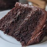 Close-up of a rich, dark slice of chocolate cake recipe with thick, fudgy frosting on a white plate.