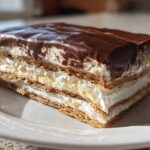 A close-up of a layered slice of chocolate eclair cake showing graham crackers, cream, and chocolate frosting.