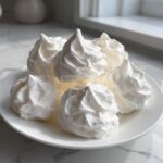 A close-up of several perfectly piped, bright white meringue cookies stacked on a white plate.