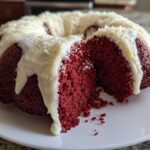 A slice removed from a vibrant red velvet bundt cake topped with thick white cream cheese drizzle.