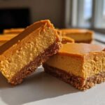 Two rich pumpkin cheesecake bars showing a thick, creamy orange filling and a graham cracker crust, ready to eat.