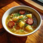 A close-up of a bowl of rich sauerkraut soup featuring chunks of potato, slices of browned sausage, and fresh dill.
