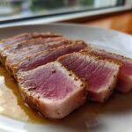 Four thick slices of perfectly seared tuna steak, showing a bright pink rare center and seasoned crust, resting in a light sauce.