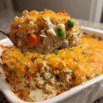 A spoonful of creamy chicken and wild rice casserole topped with melted cheddar cheese being lifted from the baking dish.