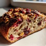 A close-up slice of homemade cranberry walnut bread showing its open crumb, topped with dried cranberries and walnuts.