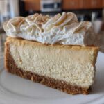 A thick slice of creamy eggnog cheesecake recipe on a white plate, topped with piped whipped cream and cinnamon.