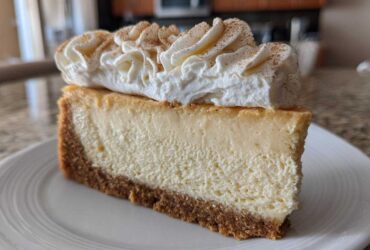 A thick slice of creamy eggnog cheesecake recipe on a white plate, topped with piped whipped cream and cinnamon.