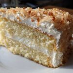 Close-up of a moist slice of 3-layer coconut cake with white frosting and toasted coconut flakes.