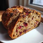 A thick slice of homemade cranberry walnut bread showing moist crumb, bright red cranberries, and walnuts, set against a white plate.