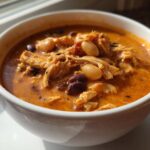 Close-up of a white bowl filled with creamy, orange-red slow cooker buffalo chicken chili featuring shredded chicken and beans.